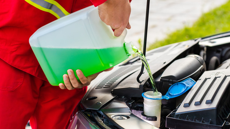 A service worker in a red uniform fills a windshield washer reservoir with green fluid from a plastic container.