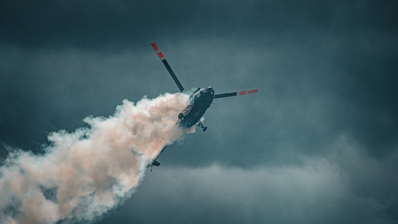 A helicopter performs an aerial stunt, trailing smoke against a cloudy sky