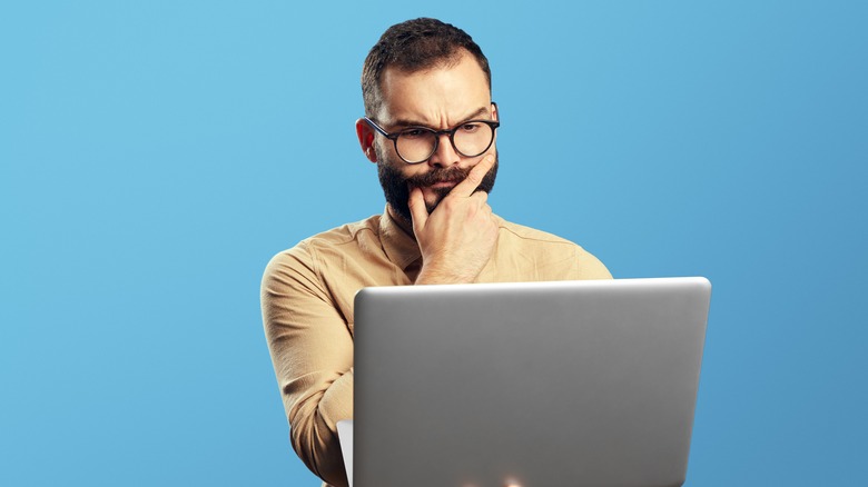 Doubtful man standng in front of blue background holding laptop
