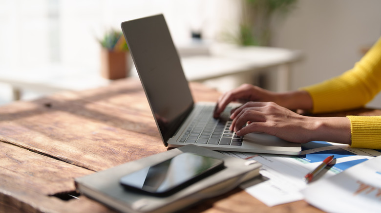 A person typing on a silver laptop on a wooden desk, with a smartphone resting on a notebook and printed charts nearby