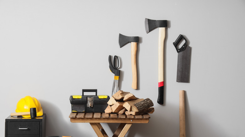 A large axe and a hatchet on a wall alongside a saw and scissors, in a room with a helmet and other tools