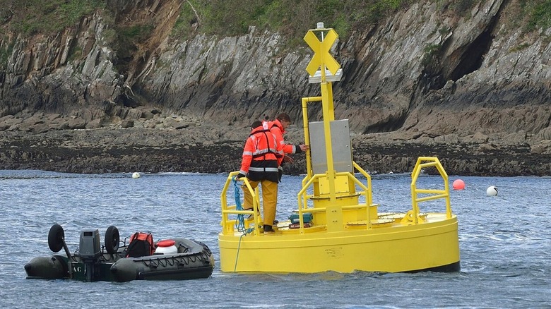 Two men in orange vests stand on large wave buoy in the water