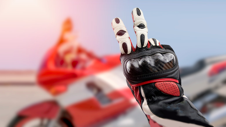 A biker wearing gloves using v sign with bike parked behind