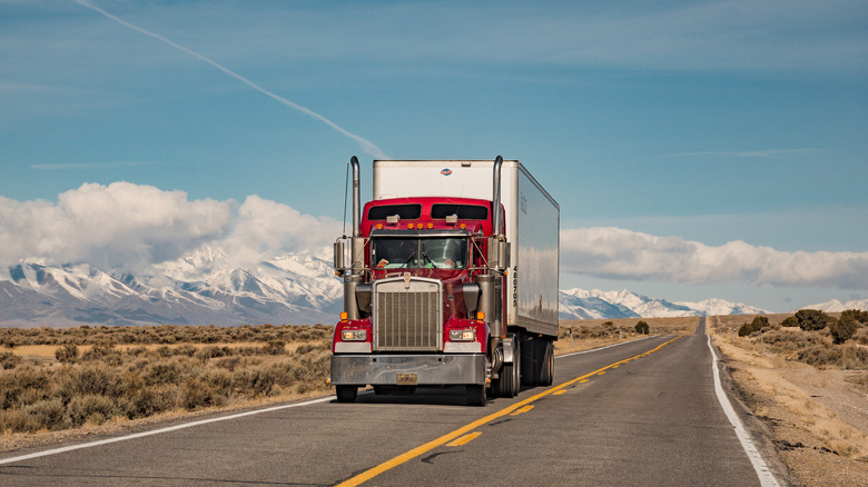 A front end shot of a red Kenworth truck driving along the highway with mountains in the background.