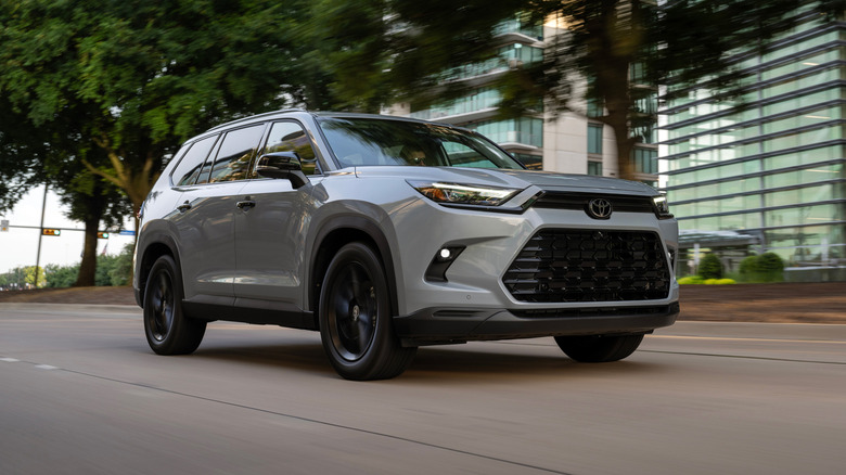 Gray 2025 Toyota Grand Highlander Nightshade driving down city street with trees and buildings in the background