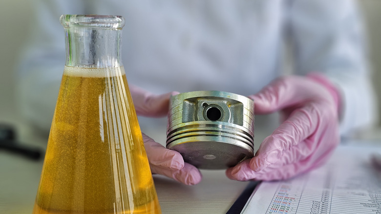 Close-up of beaker with yellow oil next to person with gloved hands holding engine piston