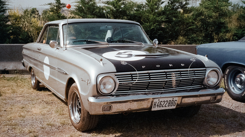An old Ford muscle car with a black hood with hood pins parked on grass.