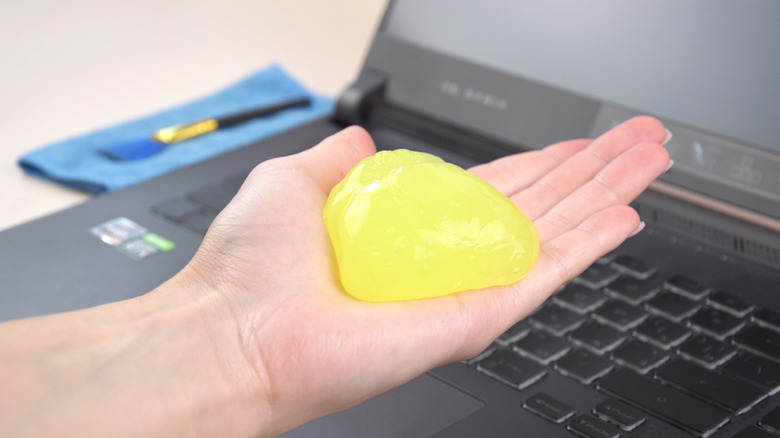 Hand holding cleaning gel with laptop opened in the background next to microfiber cloth and brush