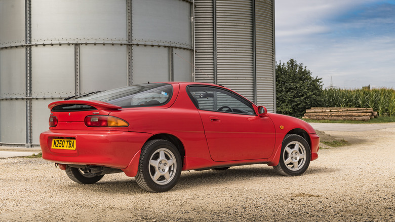 A pristine red Mazda MX-3 parked next to some metal work, rear 3/4 view