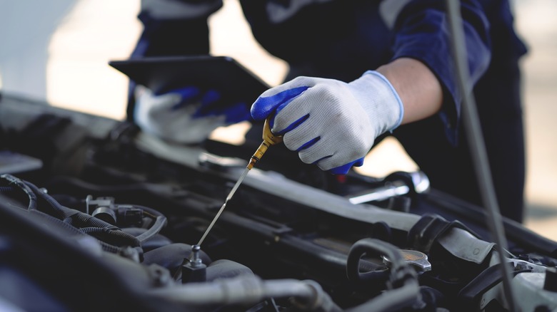 A mechanic in gloves checking an engine dipstick while holding a tablet