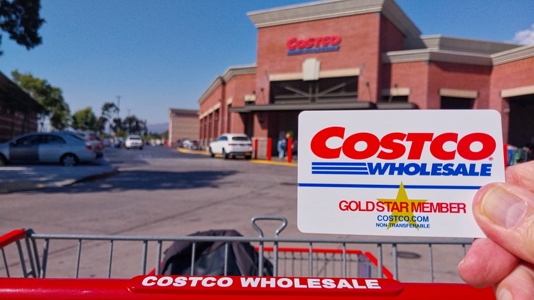 Close up of a person holding a Costco membership card with a Costco Warehouse in the backgroun