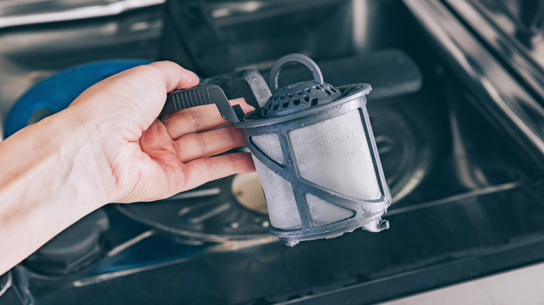 A hand holding up a dishwasher filter near an open dishwasher with a stainless steel interior