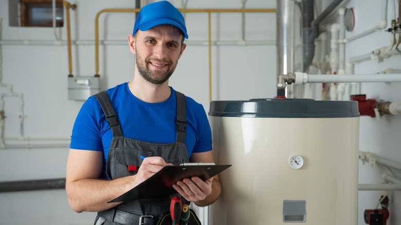A person dressed in blue overalls, holding a clipboard and inspecting a water heater