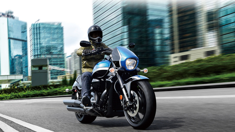 Helmeted rider on a blue Suzuki Boulevard M109R motorcycle with tall glass buildings in the background