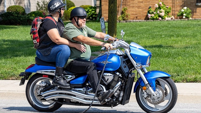 A male rider and female passenger ride a Suzuki Boulevard M109R on paved road, green grass in background