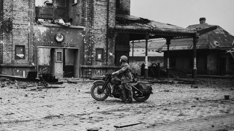 A soldier looks at the damaged Duisberg Post Office on a Harley WLA