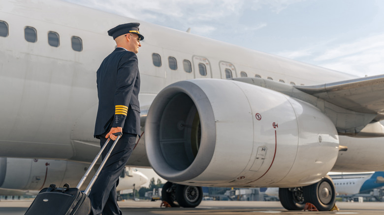 A pilot in a dark suit and hat with a suitcase next to a large white jet.