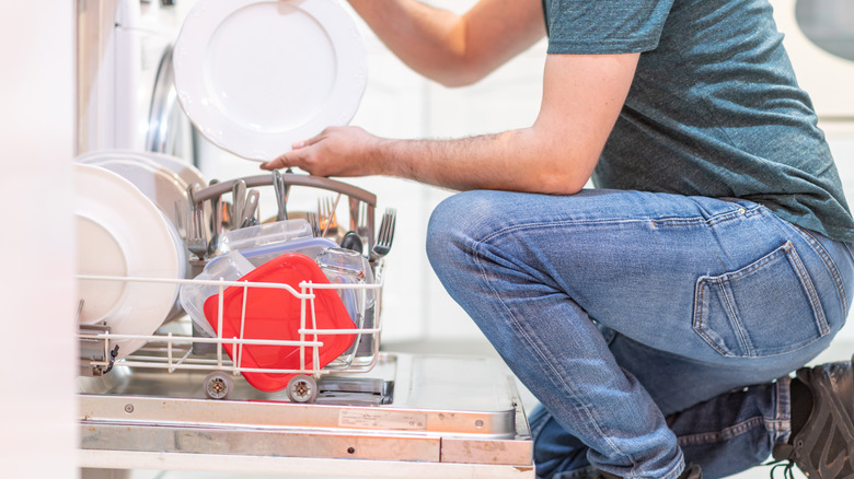 A person in blue jeans crouching to place a white plate in a loaded dishwasher.