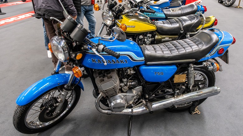 A side view of a blue Kawasaki 750 Mach IV standing first in a row of parked motorcycles.