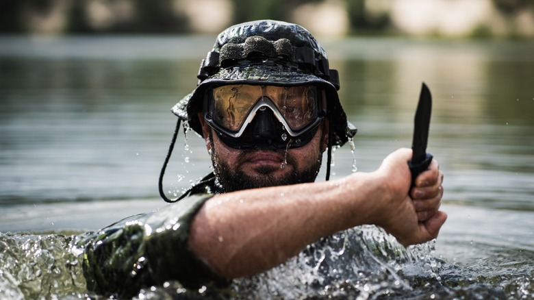 Bearded man in tactical gear and armor holding a knife while rising from the water