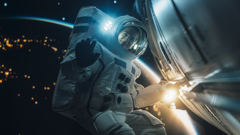 Portrait of a Female Astronaut Posing in a Space Suit During a Spacewalk Mission on a Spaceship, Satellite or a Space Station. Adult Woman Looking at Camera, Smiling and Waving Hello Gesture with Hand
