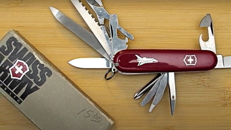 A Shuttle design Master Craftsman Victorinox Swiss Army Knife on a wooden table. All the tools are unfolded displaying blades, scissor, can opener and more. Next to the knife is the original box, which displays the words Swiss Army in large letters.
