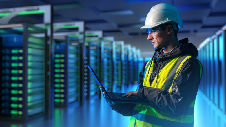 Man wearing a hard hat and high-vis vest on a laptop inside a datacenter with racks of servers in the background