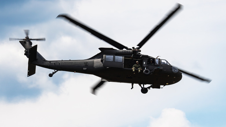 A flying UH-60M Black Hawk helicopter with a soldier sitting in the open doorway