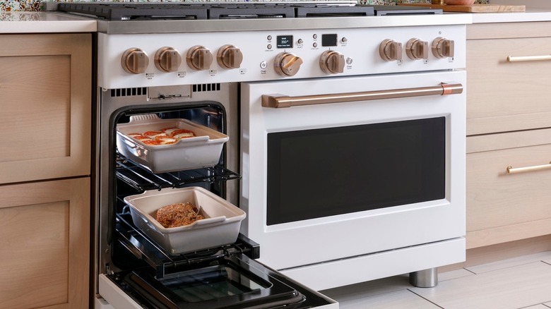 a white Café 48-inch Smart Duel-Fuel Commercial Style Range with the smaller second oven opened to reveal two large baking dishes with food in them