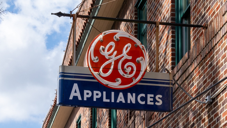 A vintage red and black GE appliances neon sign hanging outside a brick building on a sunny day