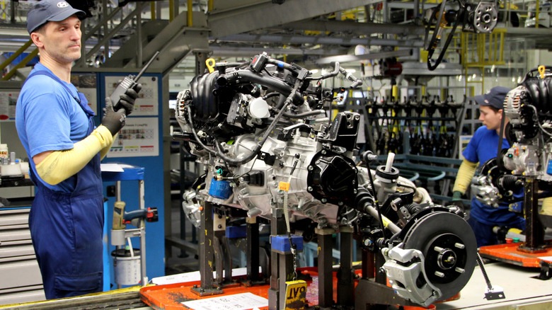 A worker in a Hyundai cap and overalls inspects an engine on an assembly line.