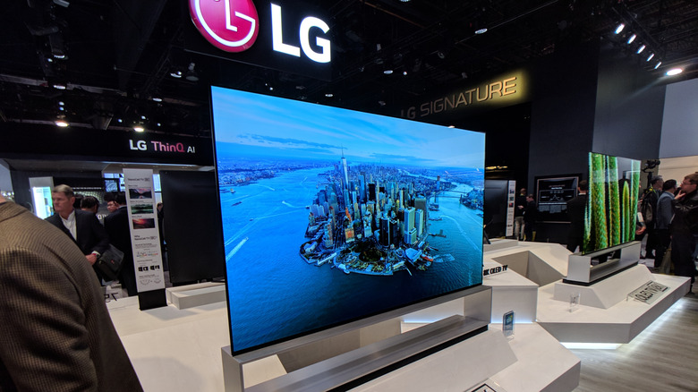LG TV on display at a trade show surrounded by other tech and people. The TV is displaying a mostly blue picture of a cityscape.