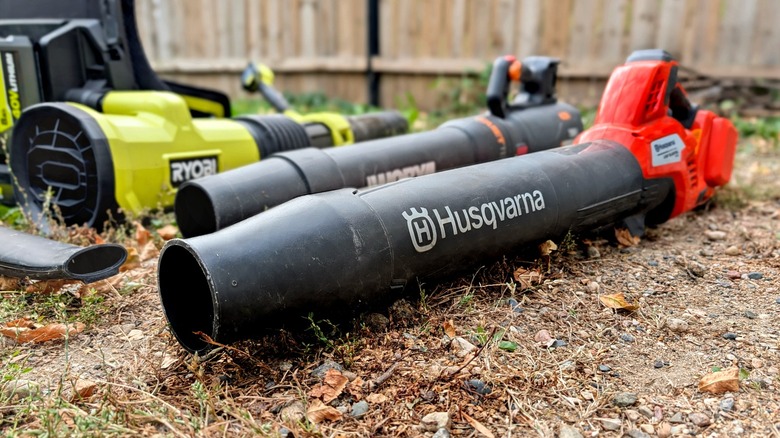 Leaf blowers lined up on the ground on gravel and grass.