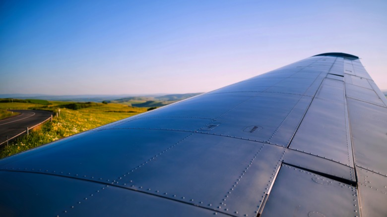 Close-up of an airplane wing, showcasing its rivets, with a rural landscape in the background