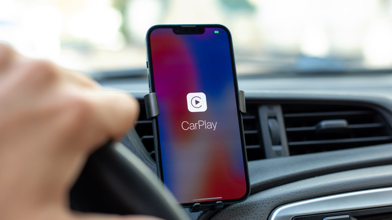 Apple CarPlay launched on an iPhone attached to a holder in a car with a driver's hand on steering visible