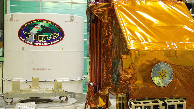 The TIRS instrument in the foreground with its shipping container waits in the background. The copper-color of TIRS is due to the gold-colored foil that coats the Multi-Layer Insulation blankets.