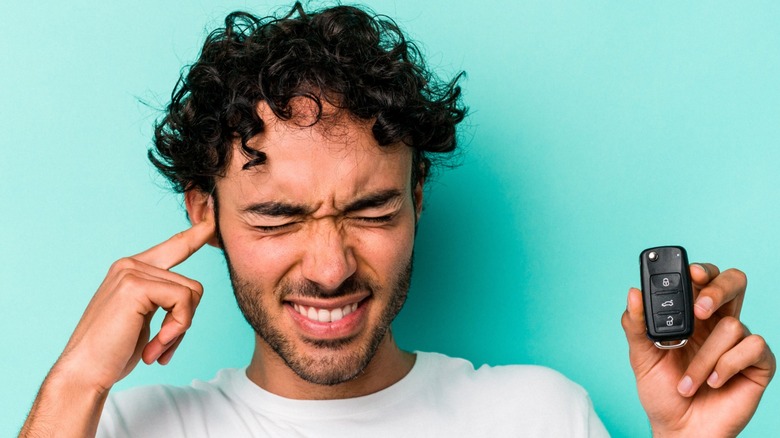 A Man Plugging One Ear And Holding A Car Key Fob In the Other With Pained Expression