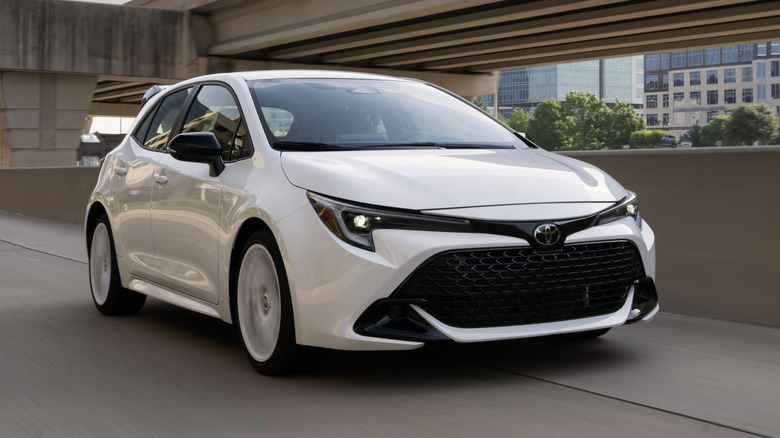White 2026 Toyota Corolla hatchback driving down a highway parallel to a tall overpass