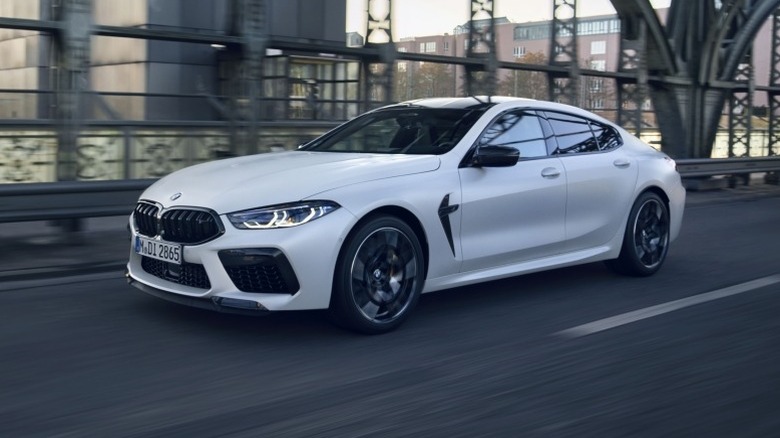 BMW M8 Competition Gran Coupe driving on an asphalt road with metal railing and buildings in the background