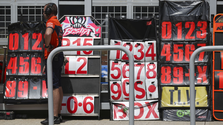 Racing engineer standing in front of four pit board number holders containing letters and numbers
