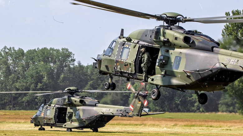 Two military helicopters with soldiers landing in grassy area