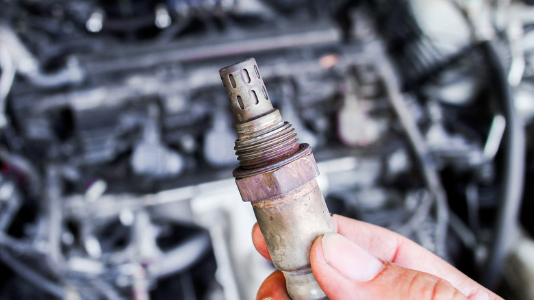 Hand holding car O2 sensor with an engine in the background