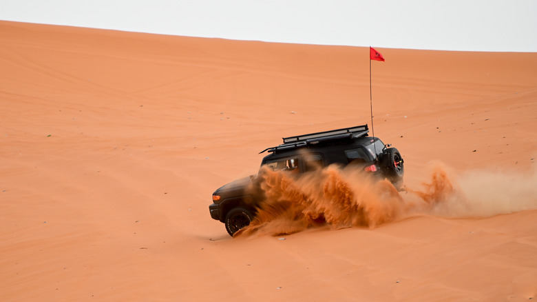 A black Toyota FJ Cruiser scaling a steep sand dune, rear 3/4 view, flag on the back
