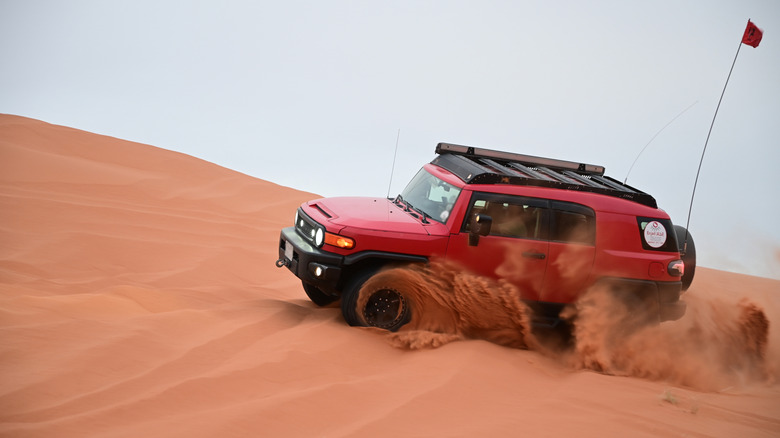 A red Toyota FJ Cruiser driving through a sand dune, front 3/4 view, flag on the back