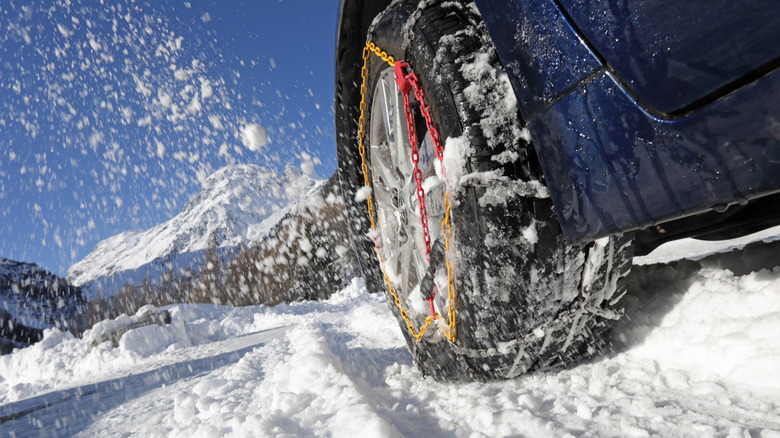 A close-up of a blue car's tire with tire chains going through snow with a mountain in the background.