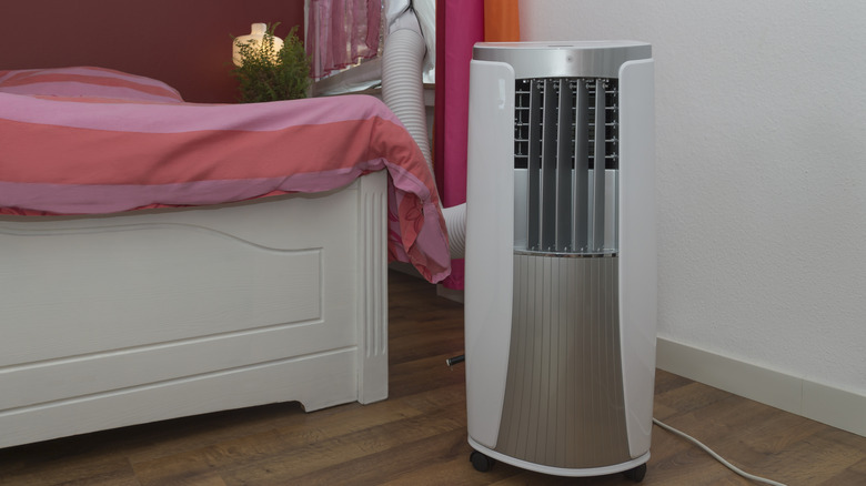 A portable air conditioner standing on the floor by a bed.
