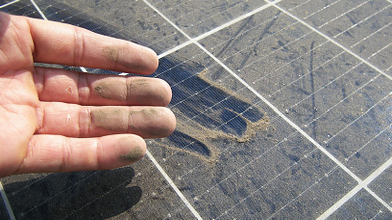 A close-up of a hand with dusty fingers from wiping them on a solar panel covered in dirt.