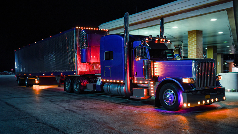 A classic semi truck with a fully chrome reefer trailer rests at the pump under the night sky