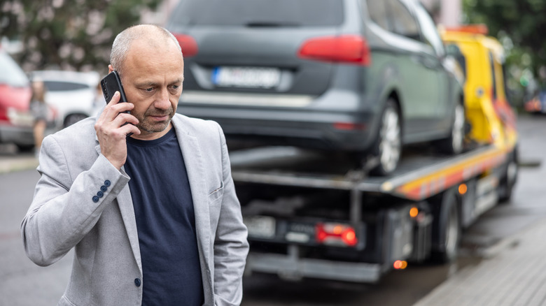 A middle-aged man calling through his phone as a tow truck carries a car at the back.