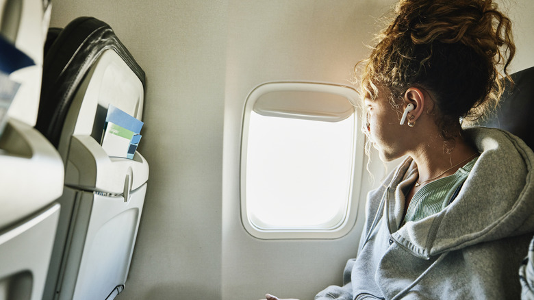 A young woman in a gray jacket and with AirPods in her ears looking out the window of commercial airplane.